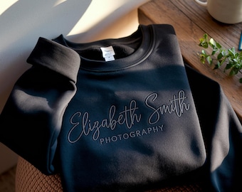 May include: A black sweatshirt with the embroidered text "Elizabeth Smith Photography" in a cursive font. The sweatshirt is folded on a brown surface, with a coffee mug and greenery in the background.