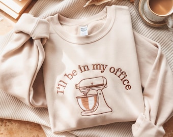 May include: Beige crewneck sweatshirt with the embroidered phrase "I'll be in my office" and a mixer graphic in brown. The sweatshirt is folded on a neutral-colored blanket, with a cup of coffee and books in the background.