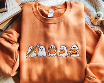 May include: Orange crewneck sweatshirt featuring embroidered white ghosts with various items: a pencil, book, coffee mug, and jack-o'-lantern. The sweatshirt is folded on a textured surface, with books and a decorative ghost visible.