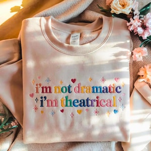 May include: A light beige sweatshirt with the embroidered text "I'm not dramatic, I'm theatrical" in colorful letters. The design includes hearts, stars, and diamond shapes. The sweatshirt is folded on a soft, textured surface.