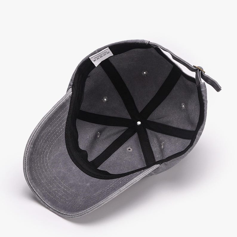 May include: A gray baseball cap with a curved brim, viewed from the inside. The cap has a black interior with a series of radiating seams. A small tag is visible inside the cap. The adjustable strap is fastened with a metal buckle.