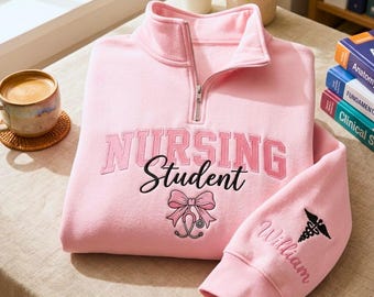 Könnte beinhalten: Hellrosa Sweatshirt mit kurzem Reißverschluss und dem gestickten Schriftzug "NURSING Student". Unter dem Text befindet sich eine Schleife und ein Stethoskop. Der Ärmel zeigt den Namen "William" und ein medizinisches Symbol, beide gestickt.