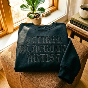 May include: A dark teal sweatshirt with the words "RETIRED BLACKOUT ARTIST" embroidered in a gothic font. The sweatshirt is folded on a brown upholstered ottoman, with a small potted plant and books visible in the background.
