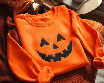 May include: A bright orange long-sleeve sweatshirt featuring a large, black, embroidered jack-o'-lantern face graphic on the front. The design includes two triangular eyes, a small triangular nose, and a wide, jagged, toothy grin.