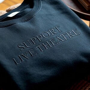 May include: Dark teal sweatshirt with the embroidered text "SUPPORT LIVE THEATRE" in a lighter shade of teal. The crew neck sweatshirt is made of a soft material.
