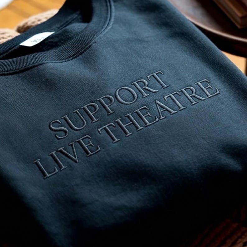 May include: Dark teal sweatshirt with the embroidered text "SUPPORT LIVE THEATRE" in a lighter shade of teal. The crew neck sweatshirt is made of a soft material.