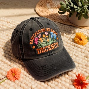 May include: A black baseball cap with the embroidered text "RADICALIZED BY BASIC DECENCY" in orange. The cap features a floral design with pink, yellow, and blue flowers. The hat is sitting on a cream-colored surface with orange flowers.