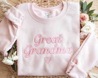 May include: A light pink sweatshirt with the embroidered text "Great Grandma" in a matching pink color, along with a small heart. The sweatshirt has long sleeves and a crew neck. The image is styled with flowers and a woven mat.