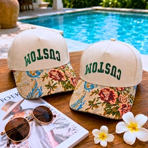May include: Two baseball caps with off-white crowns and green embroidered text reading CUSTOM. The brims feature a multi-colored floral tapestry pattern with shades of pink, blue, and gold. The caps are positioned on a wooden surface.
