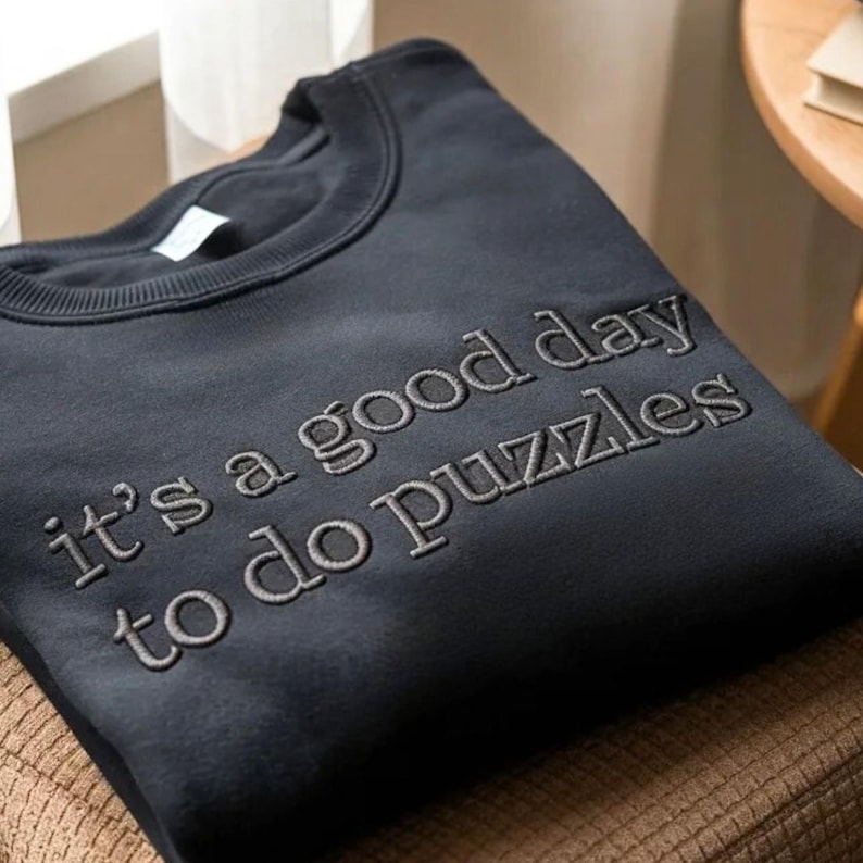 May include: A dark navy blue sweatshirt with the embroidered text "it's a good day to do puzzles" in a lighter gray thread. The sweatshirt is folded and resting on a brown surface.