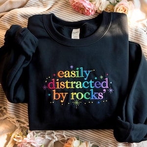 Puede incluir: Sudadera negra con el texto bordado "easily distracted by rocks" en colores arcoíris. La sudadera está doblada sobre una superficie beige a rayas, con flores rosas en el fondo.