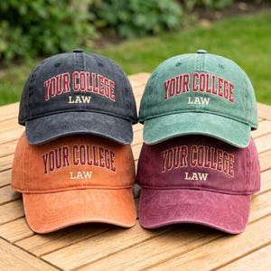 May include: Four baseball caps in various colors: black, green, orange, and burgundy. Each cap features the text "YOUR COLLEGE LAW" in a contrasting color. The caps are stacked on top of each other.