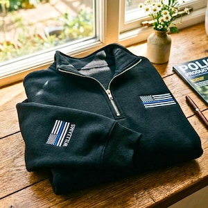May include: A black quarter-zip sweatshirt featuring an embroidered thin blue line American flag patch on the left chest and a matching flag patch with the name WILLIAMS embroidered vertically on the right sleeve cuff.