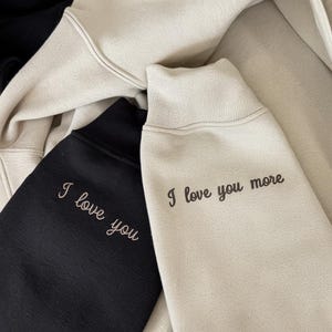 K&ouml;nnte beinhalten: Nahaufnahme von drei Sweatshirts in Schwarz und Beige. Das schwarze Sweatshirt hat den gestickten Text "I love you" in Hellrosa. Das beige Sweatshirt hat den gestickten Text "I love you more" in Dunkelbraun.