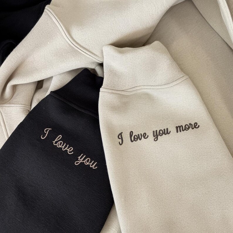 K&ouml;nnte beinhalten: Nahaufnahme von drei Sweatshirts in Schwarz und Beige. Das schwarze Sweatshirt hat den gestickten Text "I love you" in Hellrosa. Das beige Sweatshirt hat den gestickten Text "I love you more" in Dunkelbraun.