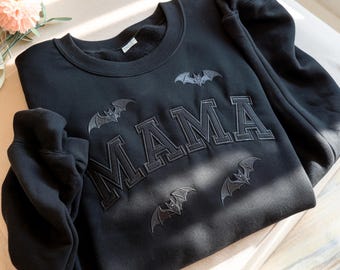 May include: A black long-sleeve sweatshirt featuring the word MAMA embroidered in a large collegiate font across the chest. The design includes four small, dark embroidered bat motifs positioned around the lettering on the front of the garment.