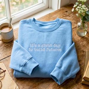Puede incluir: Una sudadera azul claro de cuello redondo con texto bordado en blanco en el pecho que dice: it's a great day to build futures. La prenda tiene mangas largas con puños acanalados y un cuello acanalado, presentada en un estilo plegado.