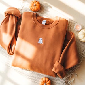 May include: A burnt orange sweatshirt with a small white ghost patch on the front. The long sleeves are folded, and the sweatshirt is surrounded by small pumpkins and a lit candle. The sweatshirt is made of a soft material.