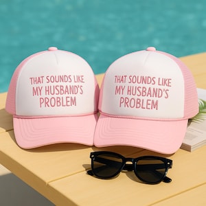 May include: Two pink and white trucker hats with the text "THAT SOUNDS LIKE MY HUSBAND'S PROBLEM" in pink lettering. Black sunglasses rest in front of the hats on a wooden surface. The hats have a mesh back and a curved brim.