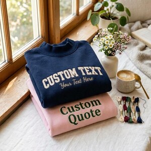 May include: A navy blue sweatshirt with the text CUSTOM TEXT Your Text Here in cream and a light pink sweatshirt with the text Custom Quote in dark green. Both sweatshirts feature a mock neck collar and are folded on a white surface.