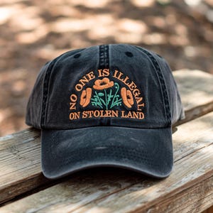 May include: Black baseball cap with the embroidered text "NO ONE IS ILLEGAL ON STOLEN LAND" in orange. The cap features an orange flower and green leaves design. The hat is sitting on a wooden surface.