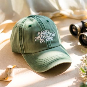 May include: A green baseball cap with the text "EASILY DISTRACTED BY BIRD" embroidered in white. The cap has a curved brim and a classic six-panel design. A small bird figurine and binoculars are in the background.