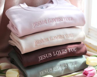 May include: A stack of four crewneck sweatshirts in pastel colors: light pink, beige, burgundy, and sage green. Each sweatshirt has the embroidered text "JESUS LOVES YOU" in a matching thread color. The sweatshirts are stacked on a pink surface.