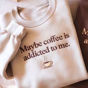May include: A beige crewneck sweatshirt featuring the dark brown text "Maybe coffee is addicted to me." centered on the chest. Below the text is a small embroidered graphic of a steaming coffee cup in shades of brown and cream.