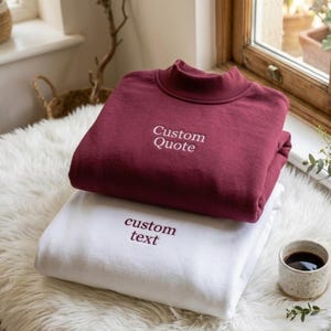 May include: A maroon mock neck sweatshirt with the white embroidered text Custom Quote stacked on top of a white sweatshirt with the maroon embroidered text custom text. Both garments feature long sleeves and a ribbed collar design.