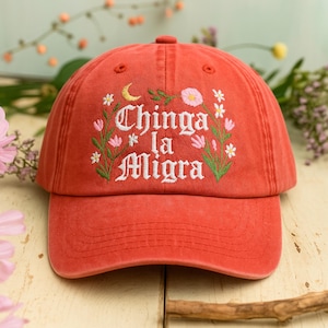 May include: A red baseball cap with the embroidered text "Chinga la Migra" in white, surrounded by floral and celestial embroidery. The cap has a curved brim and a classic six-panel design.