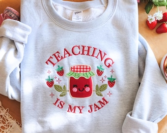 May include: A light gray sweatshirt with the embroidered phrase "TEACHING IS MY JAM" in red thread. The design features a smiling jar of jam surrounded by strawberries and leaves. A cozy, casual top.