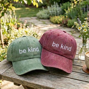 May include: Two baseball caps, one green and one burgundy, sit on a wooden table. The caps have a distressed look and feature the embroidered text "be kind" in white. The caps are a casual accessory.