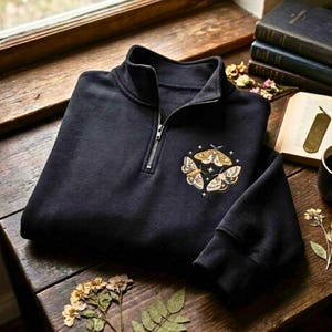 Puede incluir: Una sudadera negra con cremallera de un cuarto de largo que presenta un diseño bordado de tres polillas dispuestas en un patrón circular con pequeñas estrellas. La prenda tiene un cuello alto con tirador de metal plateado y mangas largas.