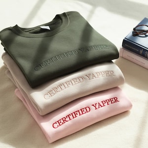 May include: Three sweatshirts in olive green, beige, and pink are stacked. Each features the embroidered text "CERTIFIED YAPPER". The sweatshirts are neatly folded, highlighting the crew neck style. The image shows a casual clothing item.