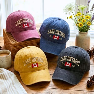 May include: Four baseball caps in maroon, blue, yellow, and charcoal gray. Each cap features embroidered white text reading LAKE ONTARIO, with EST. and 1615 flanking a small Canadian flag graphic centered on the front panel of each hat.