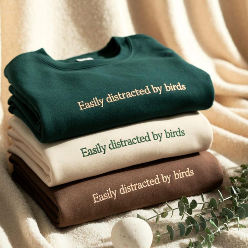 May include: A stack of three crewneck sweatshirts in dark green, cream, and brown. Each sweatshirt features the embroidered text "Easily distracted by birds" centered on the chest. The top sweatshirt has light-colored text, the middle has dark green text, and the bottom has light-colored text.