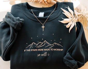 May include: A dark teal sweatshirt with the text "IF THE STARS WERE MADE TO WORSHIP so will i" and a mountain and star design. A gold cross necklace is draped over the sweatshirt. Dried flowers are in the background.