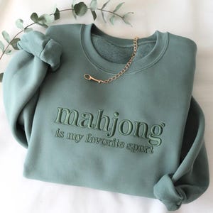May include: A sage green crewneck sweatshirt featuring the embroidered text "mahjong is my favorite sport" in a matching thread color across the chest. The garment has long sleeves with ribbed cuffs and a gold-toned chain necklace resting on the collar.