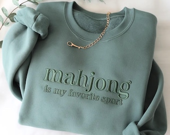 May include: A sage green crewneck sweatshirt featuring the embroidered text "mahjong is my favourite sport" in a matching thread colour across the chest. The garment has long sleeves with ribbed cuffs and a gold-toned chain necklace resting on the collar.