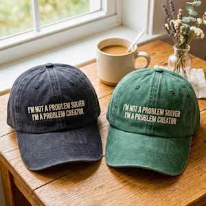 May include: Two baseball caps, one charcoal gray and one forest green, both featuring the embroidered text I'M NOT A PROBLEM SOLVER I'M A PROBLEM CREATOR in a light beige font. The caps have a distressed, washed-out fabric texture.