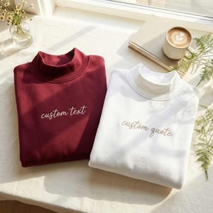 May include: A maroon sweatshirt with the embroidered text custom text and a white sweatshirt with the embroidered text custom quote, both featuring a mock neck collar and long sleeves, displayed side by side on a flat surface.