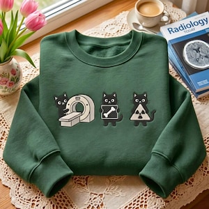 May include: A forest green crewneck sweatshirt featuring an embroidered graphic of three black cats. One cat operates a CT scanner, the second holds an X-ray of a bone, and the third holds a radiation warning symbol. The fabric is solid green.