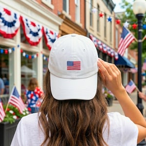 May include: A white baseball cap featuring a small, embroidered American flag centred on the front panel. The cap has a structured crown and a curved brim, constructed from a solid white fabric with matching stitching throughout the design.