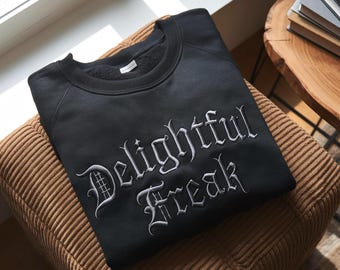 May include: A black sweatshirt featuring the embroidered words "Delightful Freak" in a gothic style. The garment is folded on a brown corduroy cushion. A window and a wooden table with books are visible in the background.