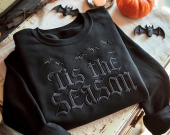 May include: A black crewneck sweatshirt featuring tonal black embroidery of the text 'Tis the Season' in a gothic font. Above the text, several small embroidered bats are arranged in a horizontal pattern across the chest of the garment.