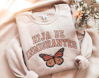 May include: A light beige sweatshirt with the text "HIJA DE INMIGRANTES" in orange lettering, and a butterfly graphic. The sweatshirt is folded on a light beige surface, with a small vase of flowers in the background.