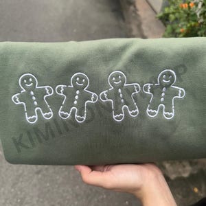 May include: A green sweatshirt with four white embroidered gingerbread men. The gingerbread men are smiling and have buttons.