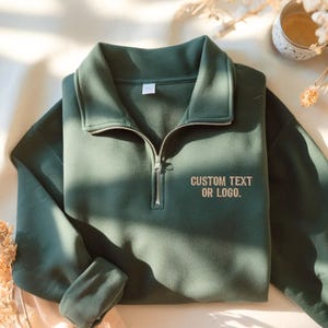 May include: A dark forest green quarter-zip sweatshirt featuring a silver-toned metal zipper and a high collar. The chest area displays the embroidered text CUSTOM TEXT OR LOGO in a neutral beige thread color, centered on the right side.
