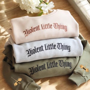 May include: Three sweatshirts stacked on top of each other. The top sweatshirt is beige with the text "Violent Little Thing" embroidered in a dark red color. The middle sweatshirt is light gray with the text "Violent Little Thing" embroidered in black. The bottom sweatshirt is olive green with the text "Violent Little Thing" embroidered in black.