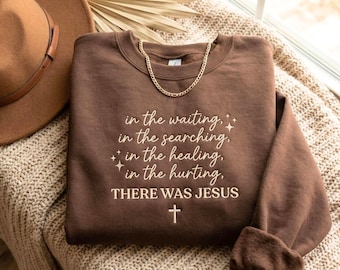 May include: A brown sweatshirt with the text "in the waiting, in the searching, in the healing, in the hurting, THERE WAS JESUS" and a cross. A gold chain is draped across the top. A brown hat is visible in the background.